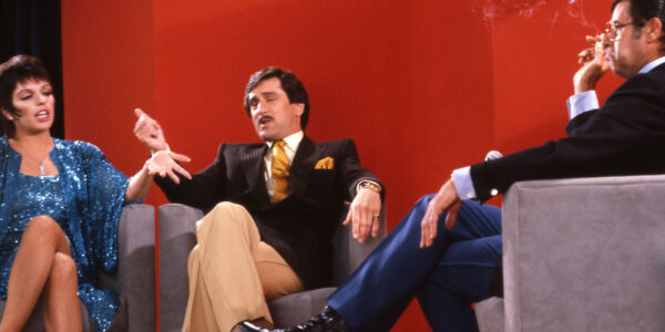 Three people sit on a talk show set with a red background