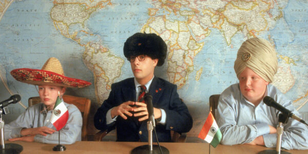 Three white students sit at a desk behind a mic and in front of a world map, the middle student wearing glasses and a Russian hat, the others wearing a turban and sombrero