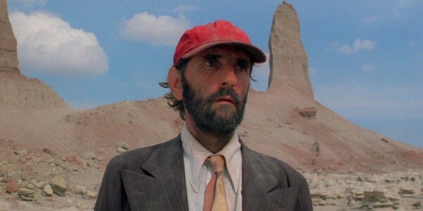 A bearded man in a red baseball cap stands in front of a sandstone tower in the desert, looking off camera