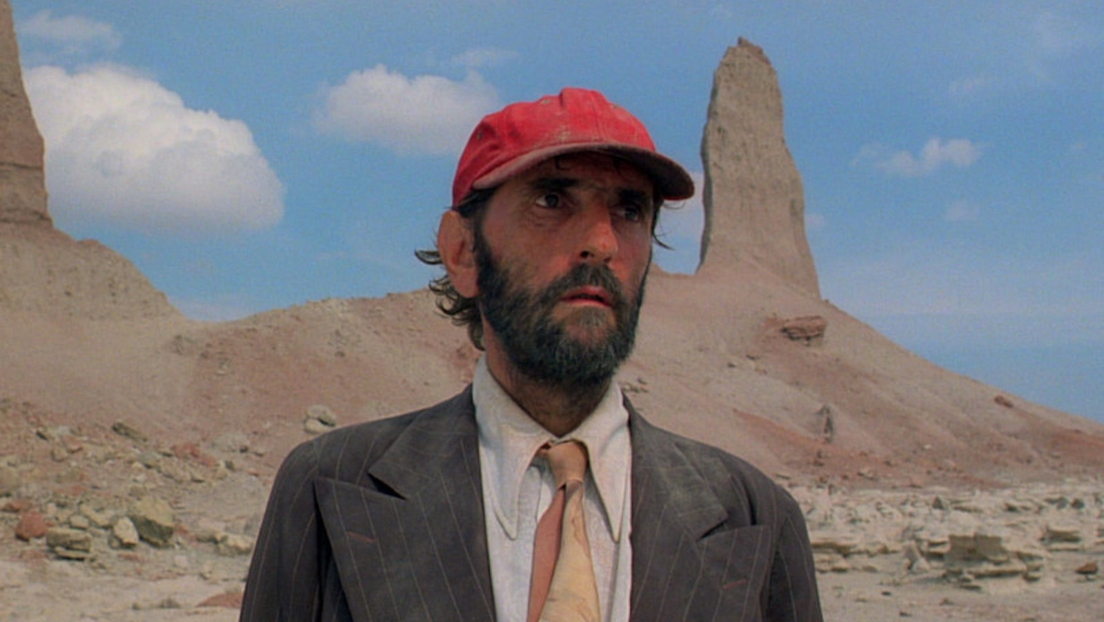 A bearded man in a red baseball cap stands in front of a sandstone tower in the desert, looking off camera