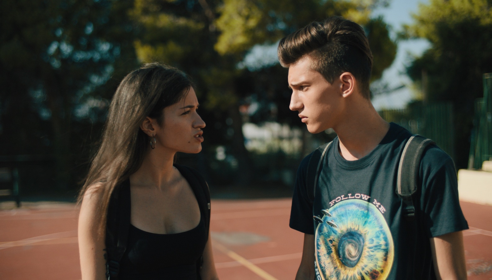 a young man and woman scowl and face each other on an outdoor basketball court
