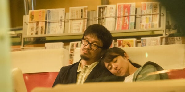 A young woman rests her head on a bespectacled man's shoulder in a bus station