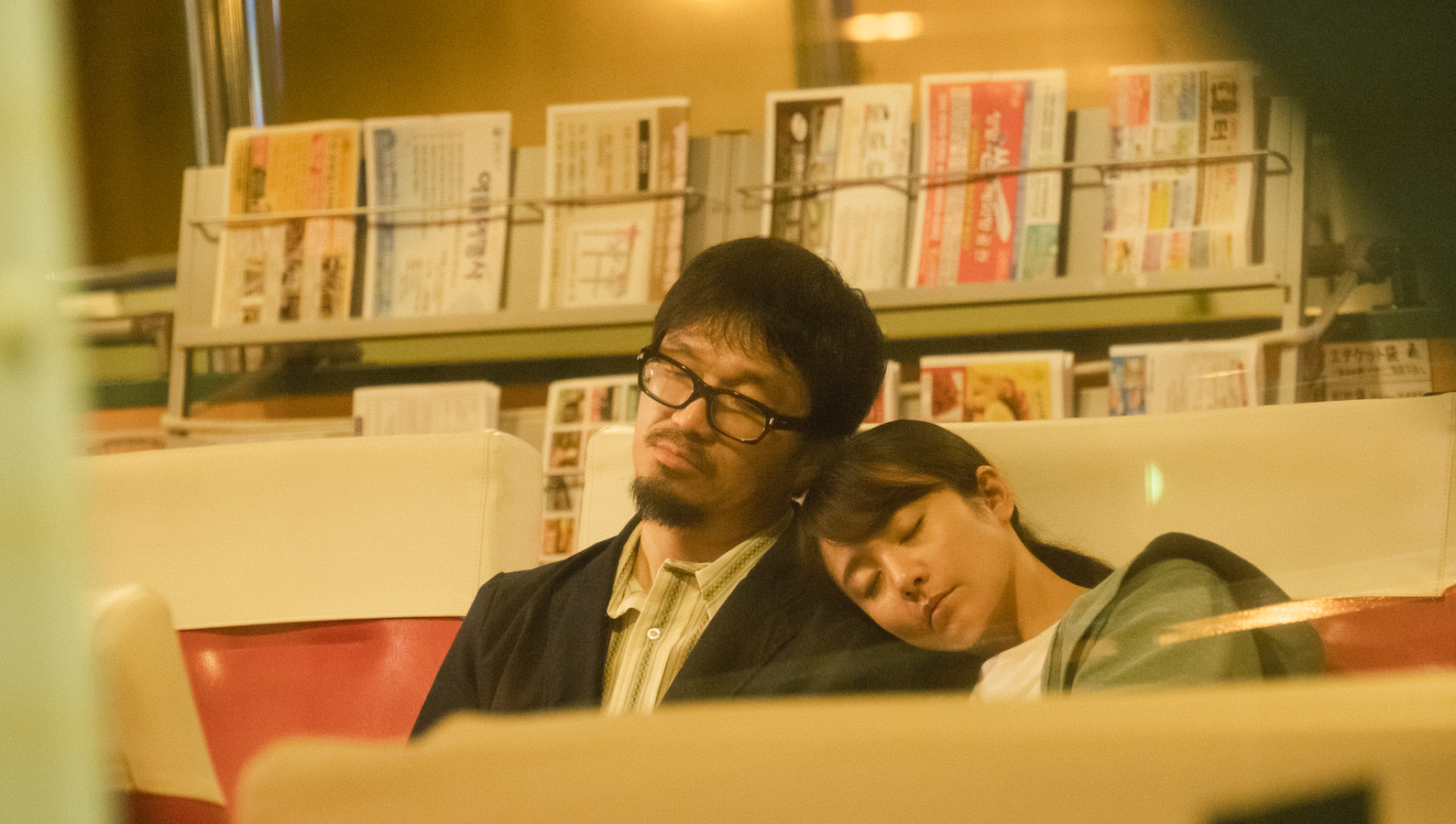 A young woman rests her head on a bespectacled man's shoulder in a bus station