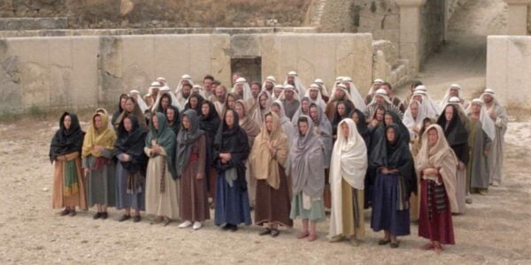 A group of people in Biblical clothing and shawls stand in a sunny desert area, lined up in single file in four rows