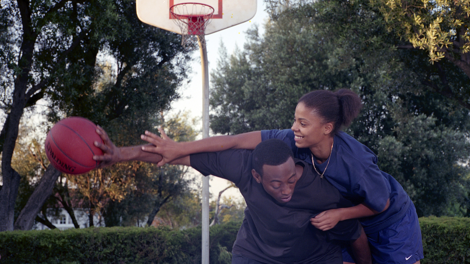 A young man and woman play basketball at an outdoor court, she's trying to block him while smiling