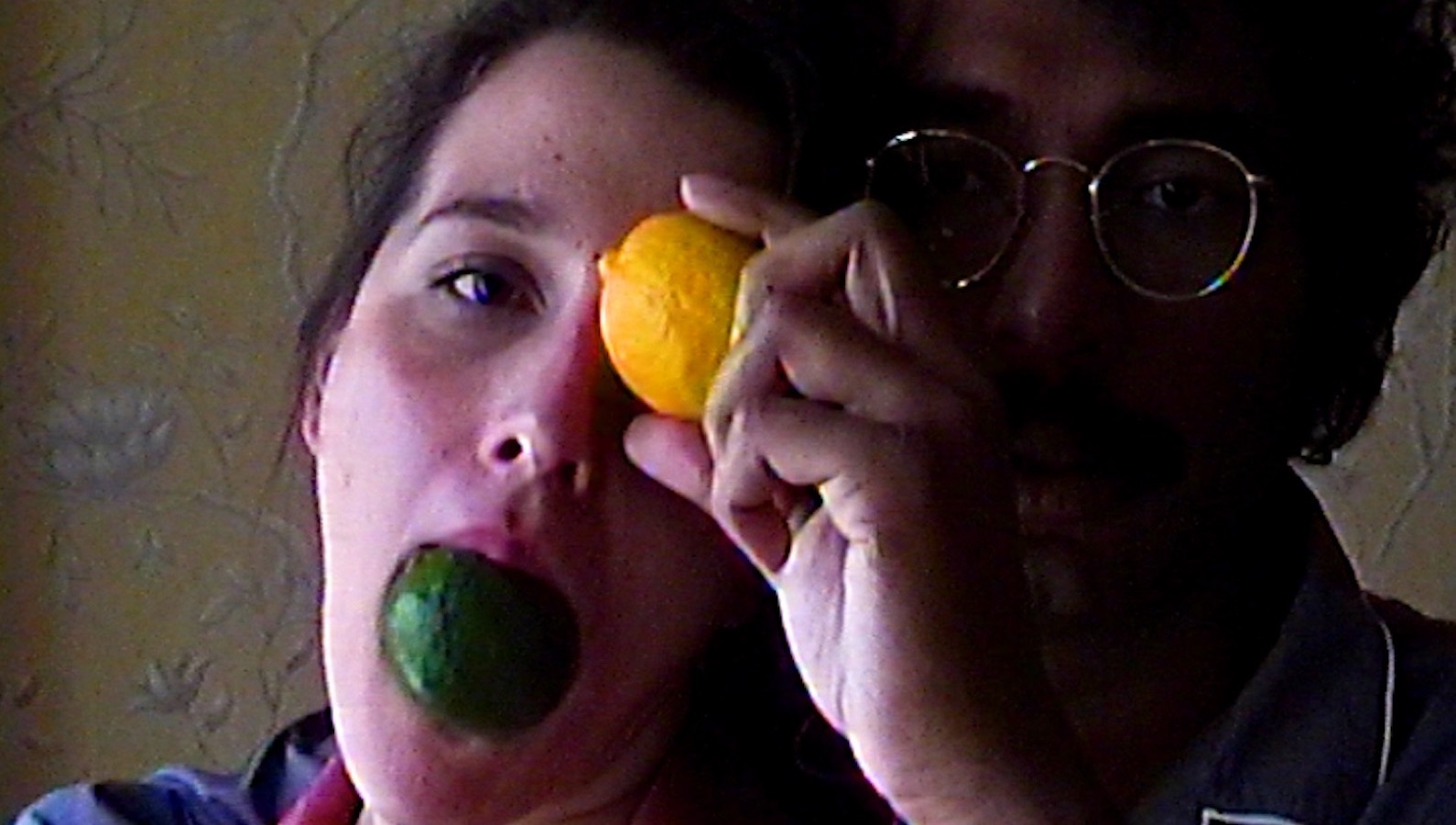 A woman holds a lime in her mouth and a lemon against her eye, with a bespectacled man behind her