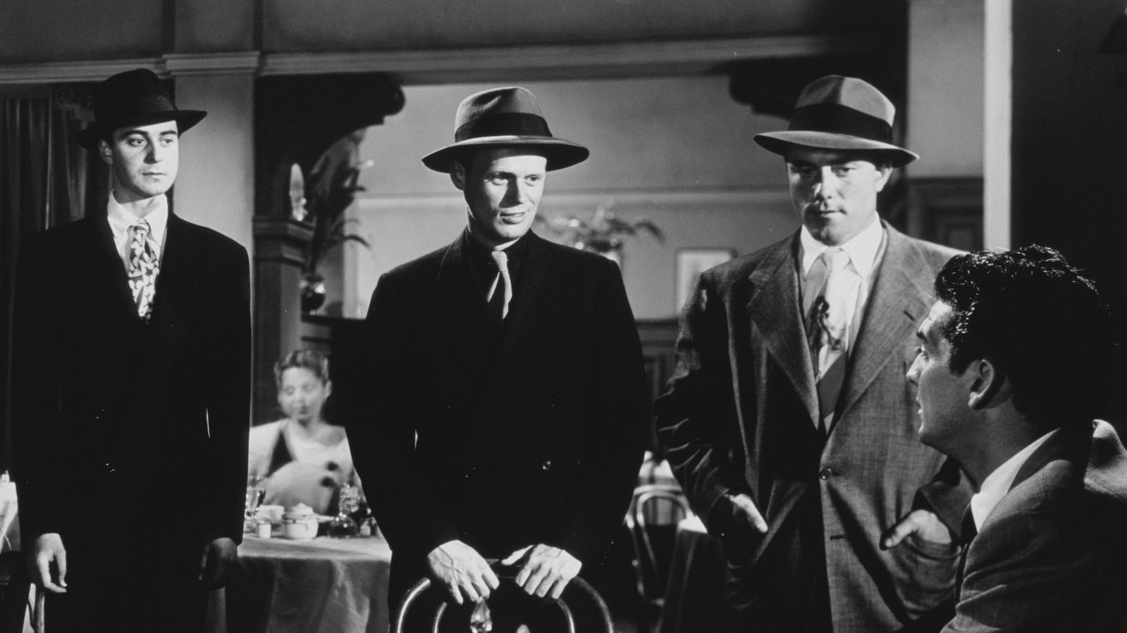 Three menacing gangsters in fedoras hover over a man sitting at a restaurant table in a black and white image