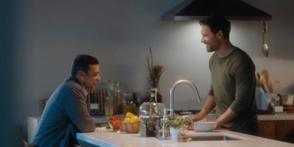 Two men in a kitchen, one sitting in front of an island, the other standing over the island, his head under a ceiling lamp