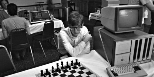 A young man in glasses sits before a chess board and an old fashioned desktop computer in a black and white image