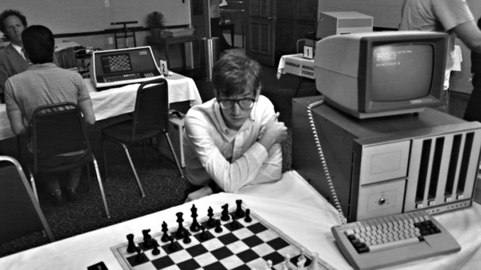 A young man in glasses sits before a chess board and an old fashioned desktop computer in a black and white image