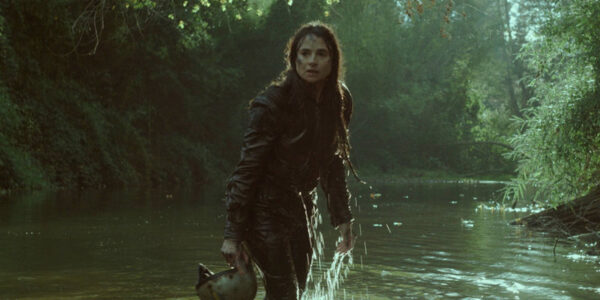 A long-haired woman carrying a helmet stands up to her knees in a river surrounded by woods