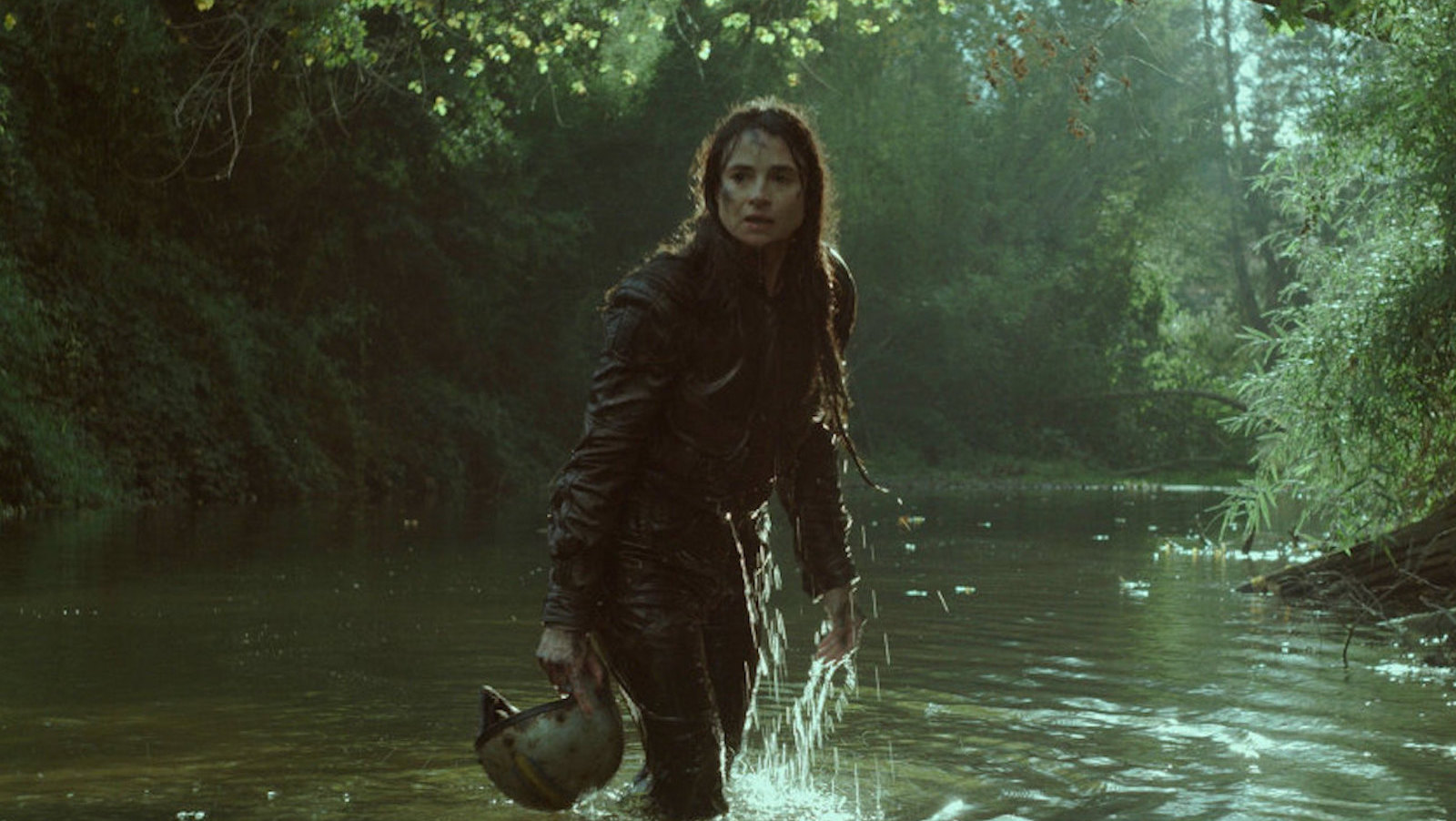 A long-haired woman carrying a helmet stands up to her knees in a river surrounded by woods