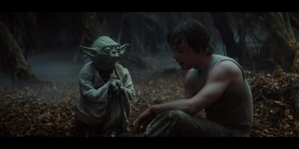 A small green creature named Yoda leans on a cane as he looks at a man in a tank top in a wooded area