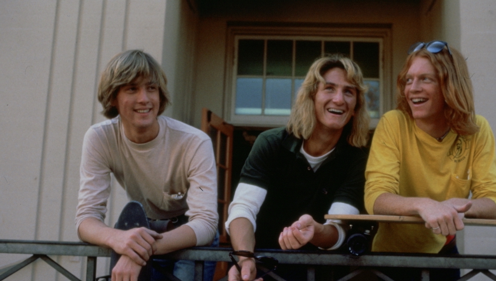 A trio of high school boys on a balcony with long blonde or red hair laugh pleasantly.