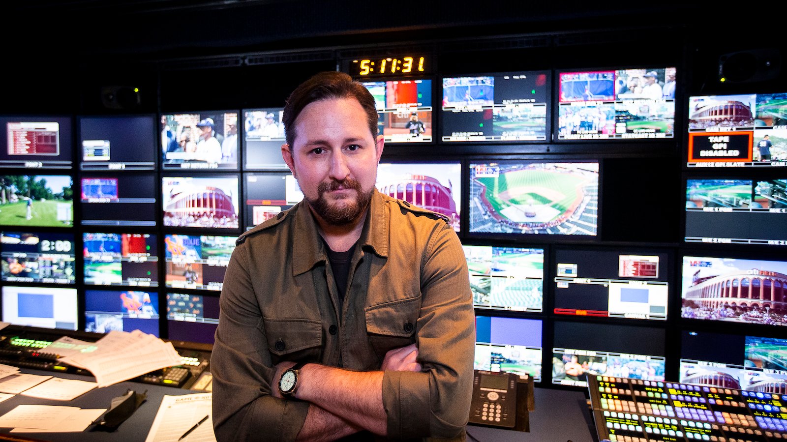 A man with a goatee stands with arms folded before a live baseball TV control room, looking at camera
