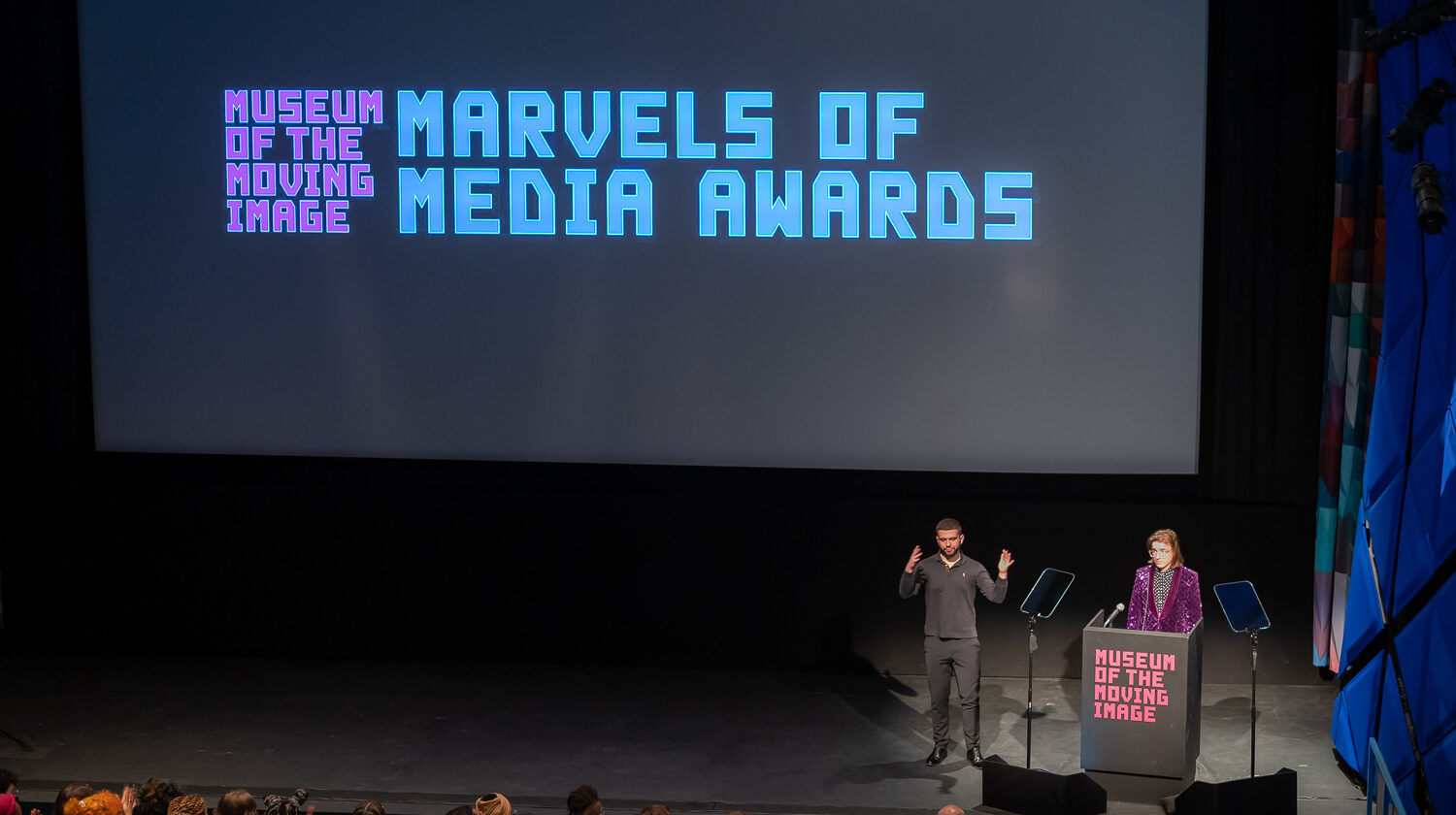 a white man in a sparkly purple suit stands at a podium on a large stage with a sign-language interpreter next to him while on the screen there are words that read Marvels of Media Awards