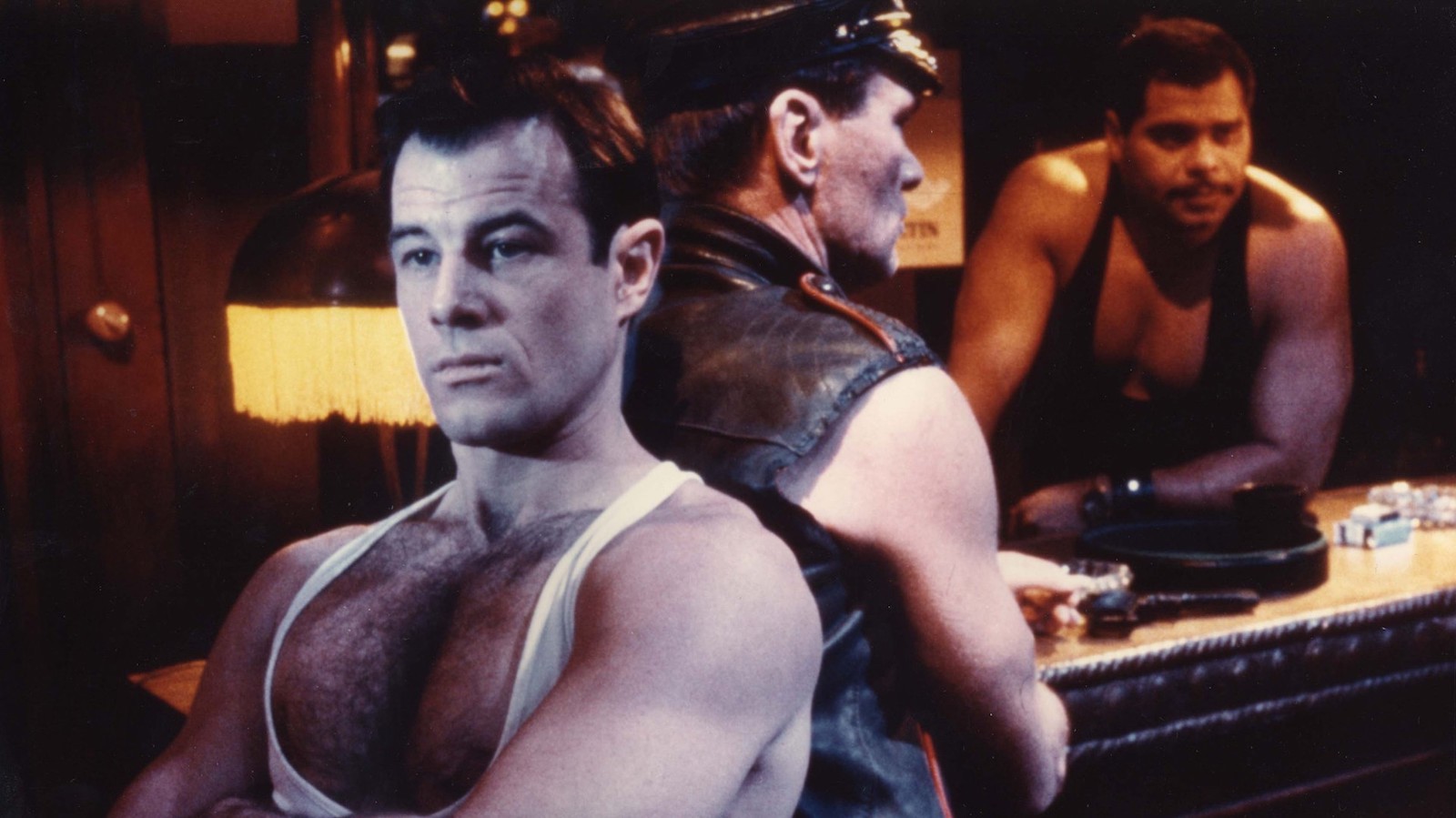 A man in a low cut tank top sits with his arms crossed, his back to another man wearing a leather cap and vest in a bar, with a bartender watching from behind them