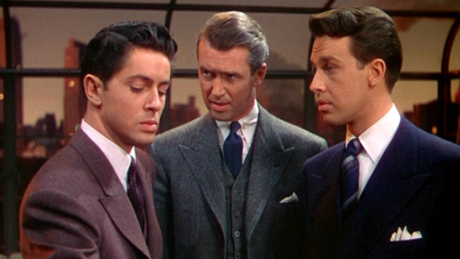 Three men stand in a room by a large window during sunset, all three wear suits, while the older man in the middle looks intently at the man on the left, who looks away painfully