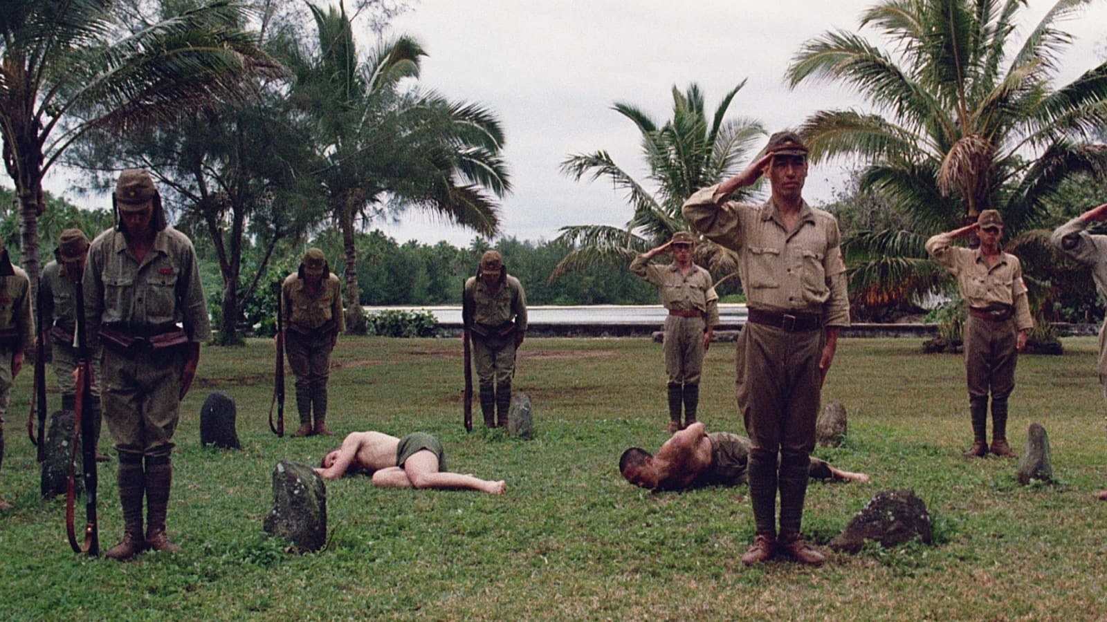 military men salute facing the camera standing amidst palm trees and two bodies writhing on the ground