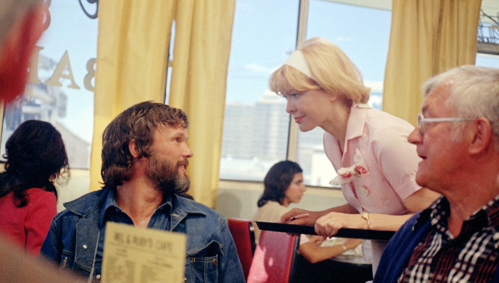 A waitress in a diner and pink shirt bends over to talk to a customer in a denim shirt