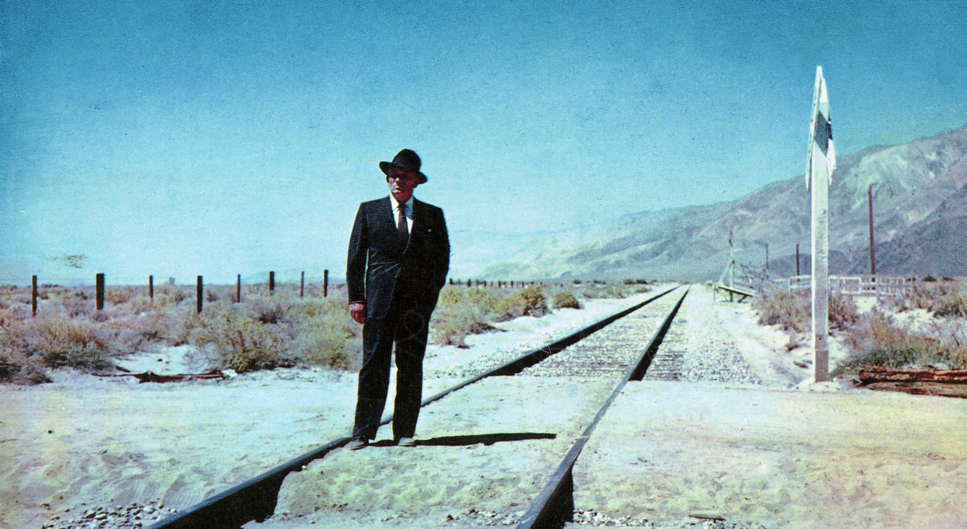 A one-armed man in a black suit and fedora stands on a railroad track in the middle of nowhere against a blue sky