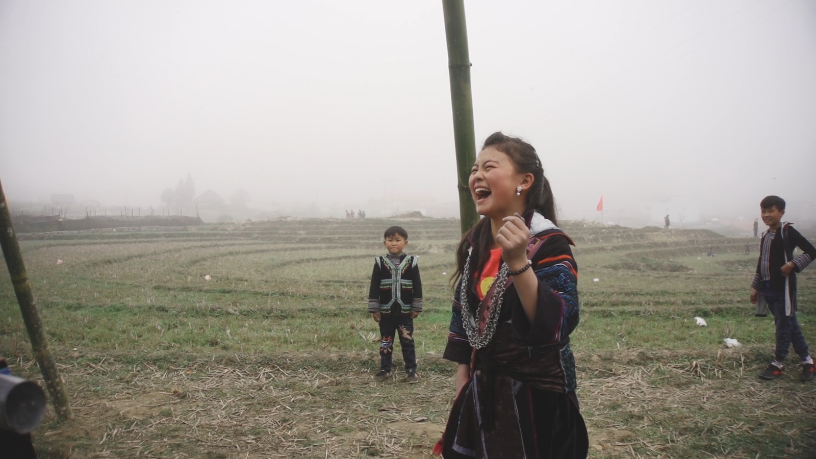 A young girl stands in a misty field and laughs joyously, with two younger boys in the background
