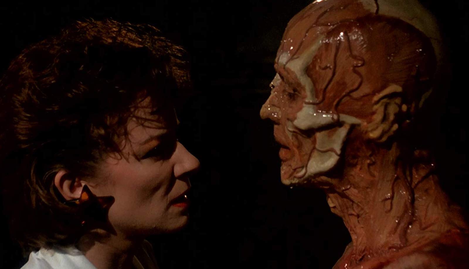 A woman and a man with a veiny, skinless face look at each other intently