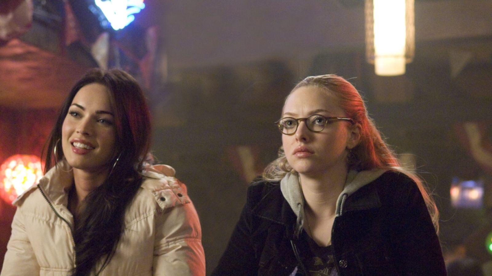 Two young women sit at a bar, one blonde, one brunette, the blonde isn't smiling, the brunette is, as they look off camera