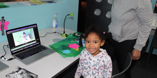 A little girl sits in front of a laptop computer with an animated princess on the screen, and she looks at the camera smiling