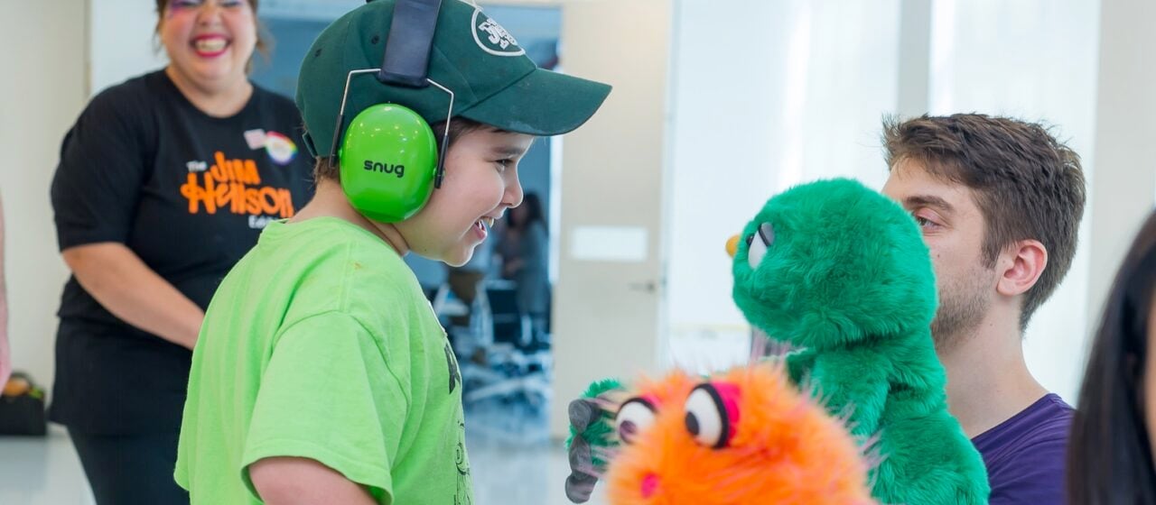 A little boy with green headphones and a green shirt interacts with a green puppet ...another orange puppet is in foreground.