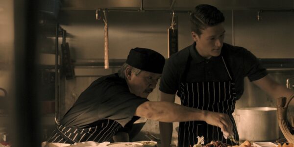 In a kitchen, an older chef reaches across a plate in front of a younger chef, whom he is instructing.