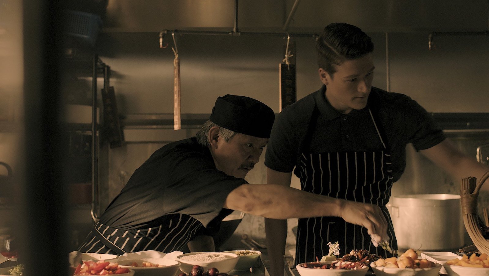 In a kitchen, an older chef reaches across a plate in front of a younger chef, whom he is instructing.