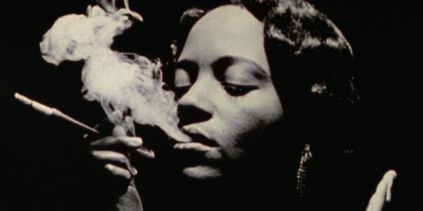 black and white image of a woman smoking, holding a cigarette holder, with her eyes closed and smoke out of her mouth like vapor