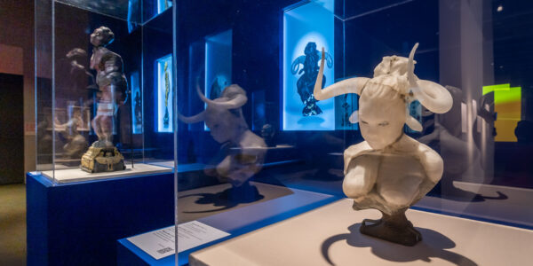 A museum gallery featuring a sculptural bust of a mythological creature—a female minotaur—with a human head and curved horns—behind a glass case.