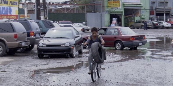 A young boy rides a bicycle through a wet parking lot with a car tire hanging from his handlebars