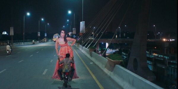 A man on a motorcycle rides across a bridge carrying a large cardboard cutout of a woman dressed in pink