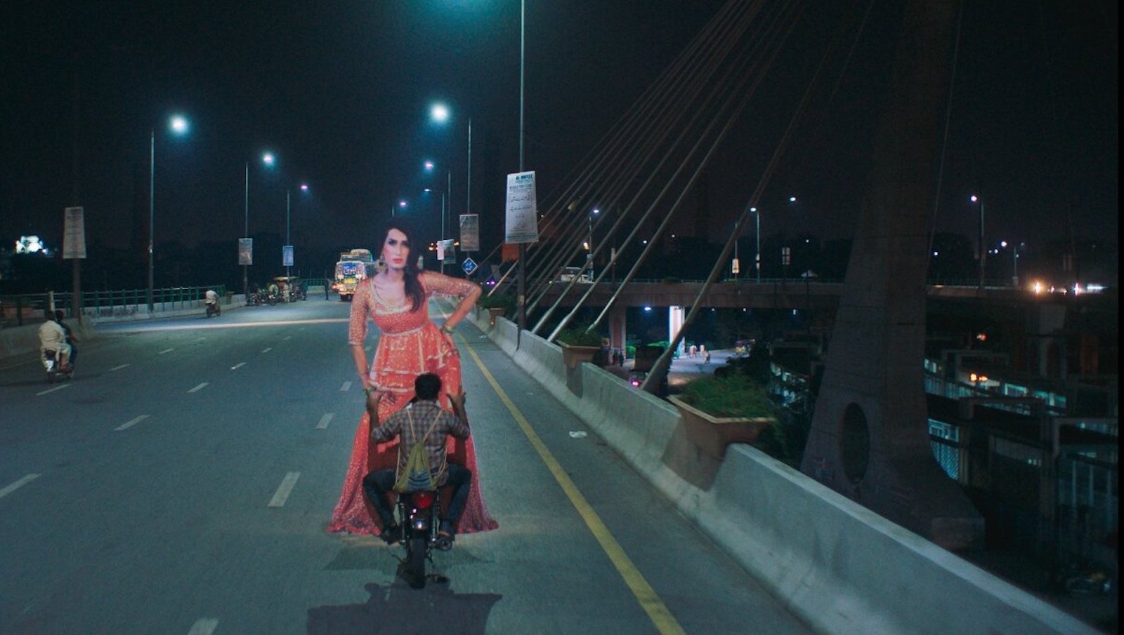 A man on a motorcycle rides across a bridge carrying a large cardboard cutout of a woman dressed in pink