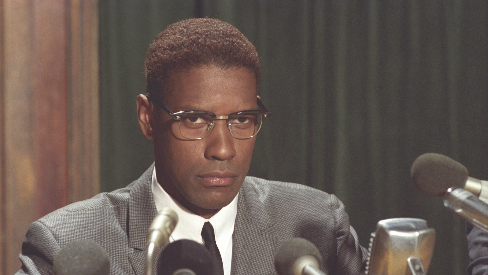 A man wearing spectacles and a serious expression sits in front of a bunch of press microphones