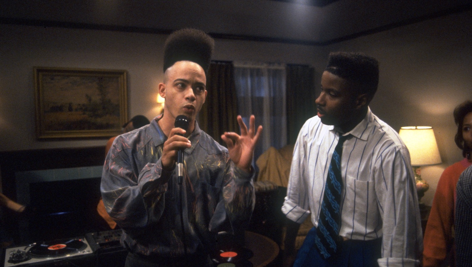A man with a tall afro hairdo stands in a living room talking into a microphone while another man, wearing a shirt and tie, looks at him curiously