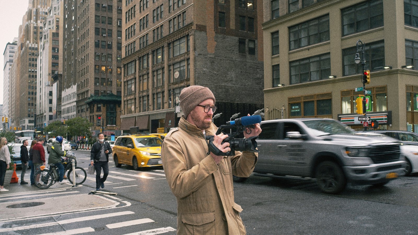 A man in glasses holds a video camera as he stands on a New York City street