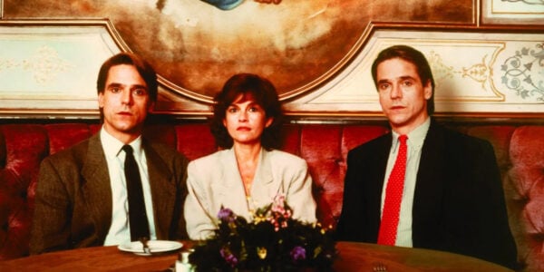 Twin brothers sit at a restaurant—one with a red tie, one with a black tie—with a woman between them, all looking at camera.