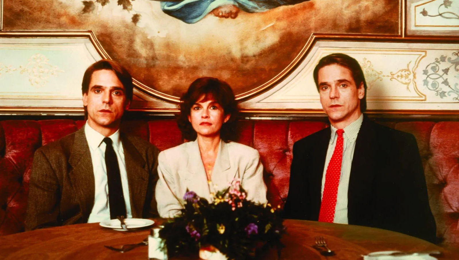 Twin brothers sit at a restaurant—one with a red tie, one with a black tie—with a woman between them, all looking at camera.
