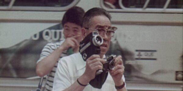 A man with flip sunglasses holds an 8mm camera and a photography camera simultaneously as he stands in front of the side of a bus, with a young man in a striped shirt behind him