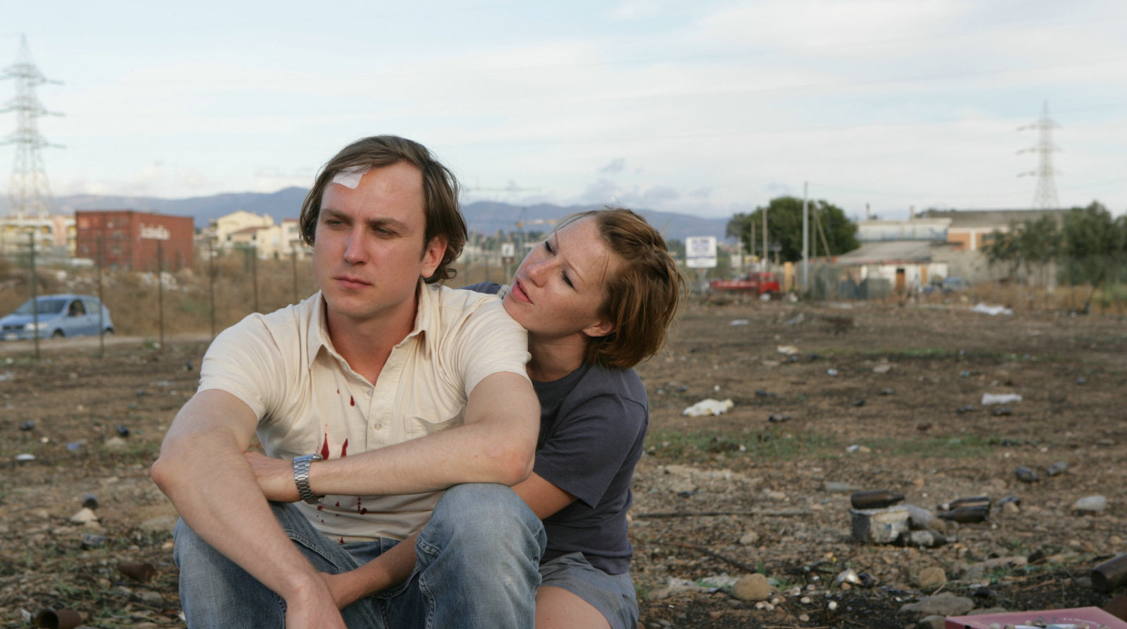 A man with a bandage on his head is embraced by a woman while sitting in a dump lot