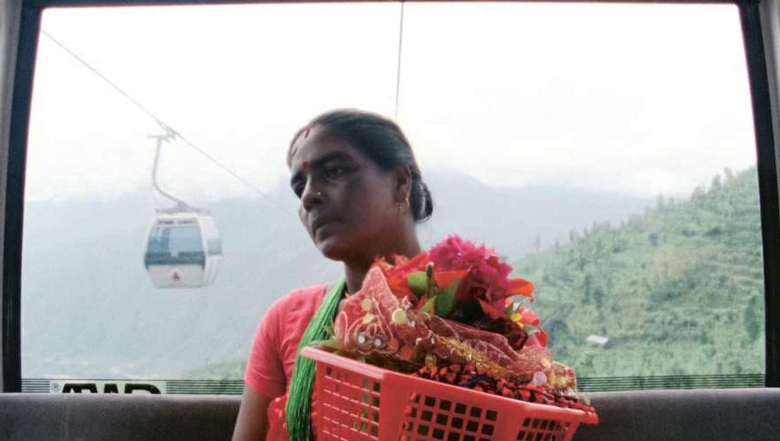 A woman carries a red basket full of flowers in a gondola high in the mountains