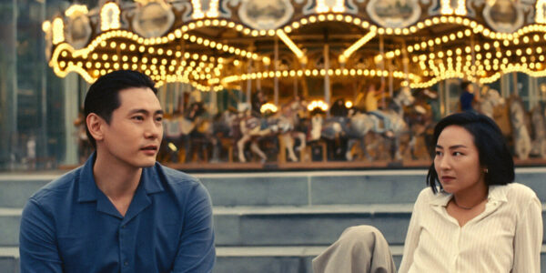 A man and woman sit on either side of the photograph, facing the camera and sitting in front of a lit-up merry-go-round.