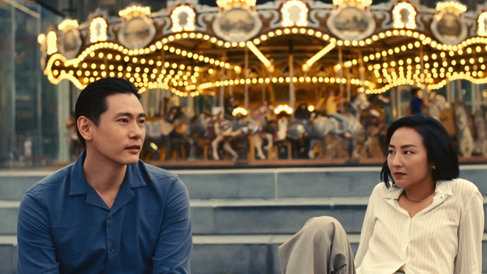 A man and woman sit on either side of the photograph, facing the camera and sitting in front of a lit-up merry-go-round.