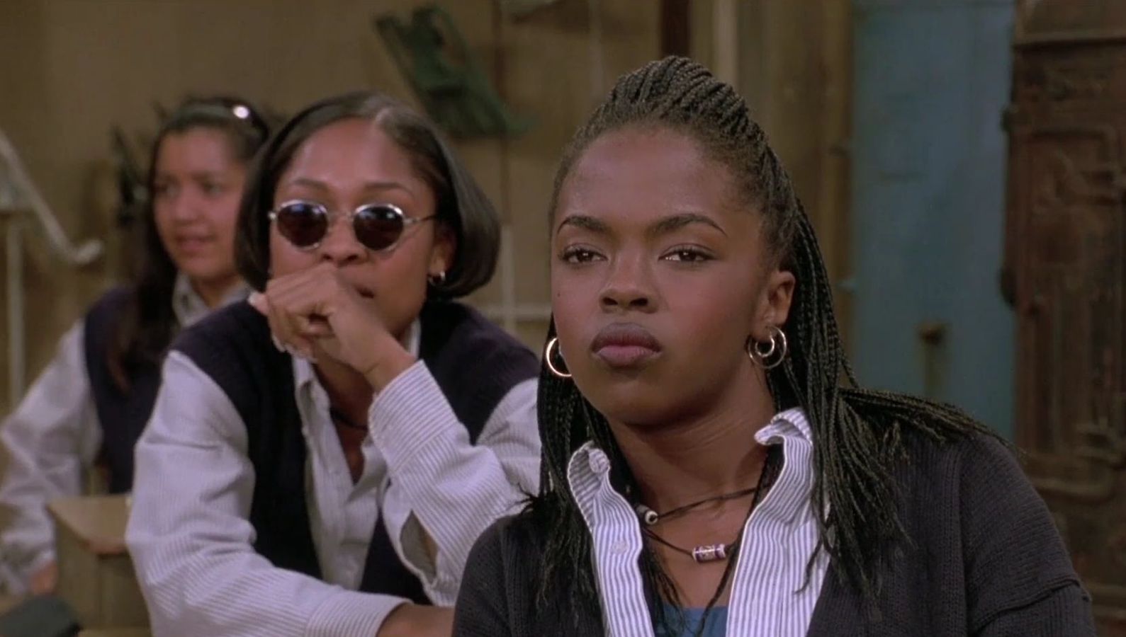 Three young women sitting in a classroom looking towards the camera; the woman in the middle-ground wearing sunglasses, the woman in the foreground staring with attitude