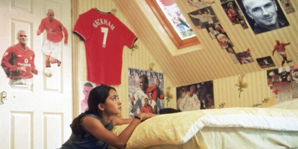 A young girl kneels by her bed and looks up at a photo of David Beckham, with images of soccer players all over her wall