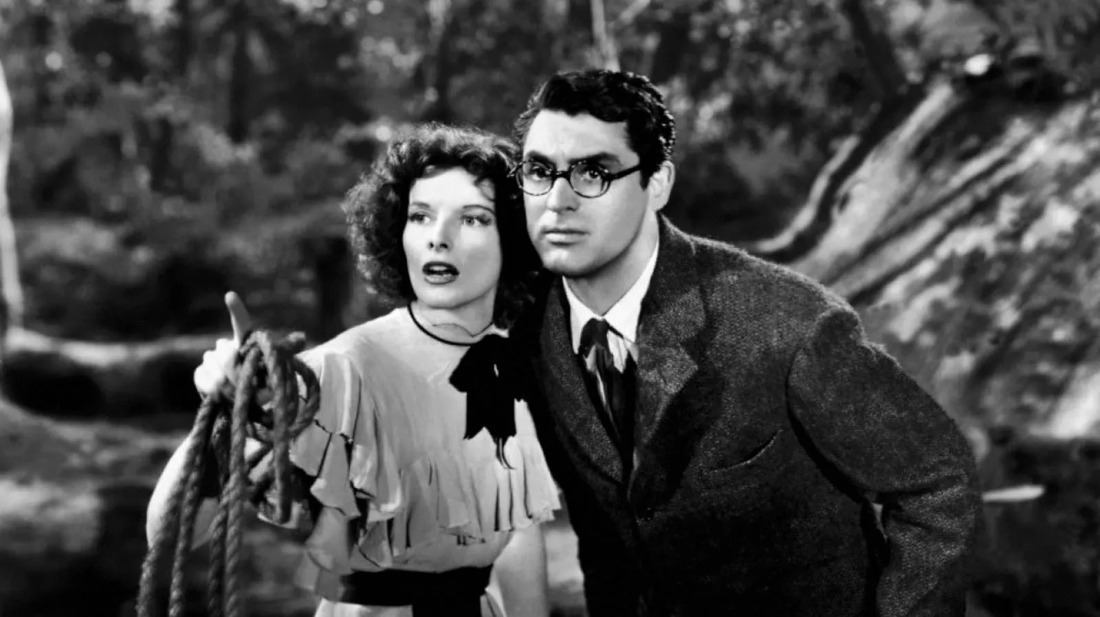 A woman holding a rope and a bespectacled man in a suit look intently off screen